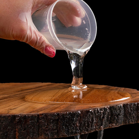 Hand pouring crystal sheen epoxy from a plastic cup onto a wooden surface with a black background