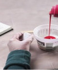 Person pouring quick mold silicone from a bottle into a white container on a light surface.