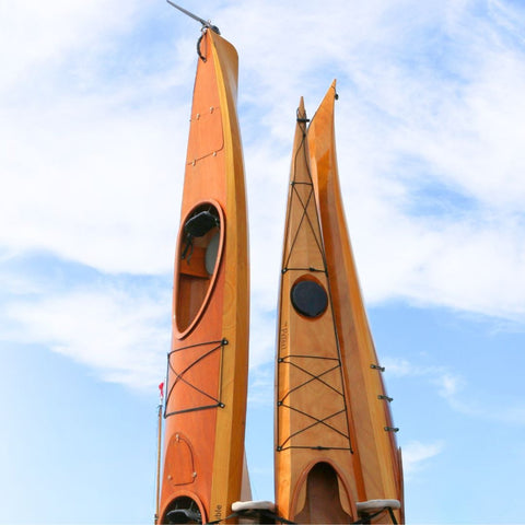 Two orange kayaks standing vertically against a blue sky with white clouds.
